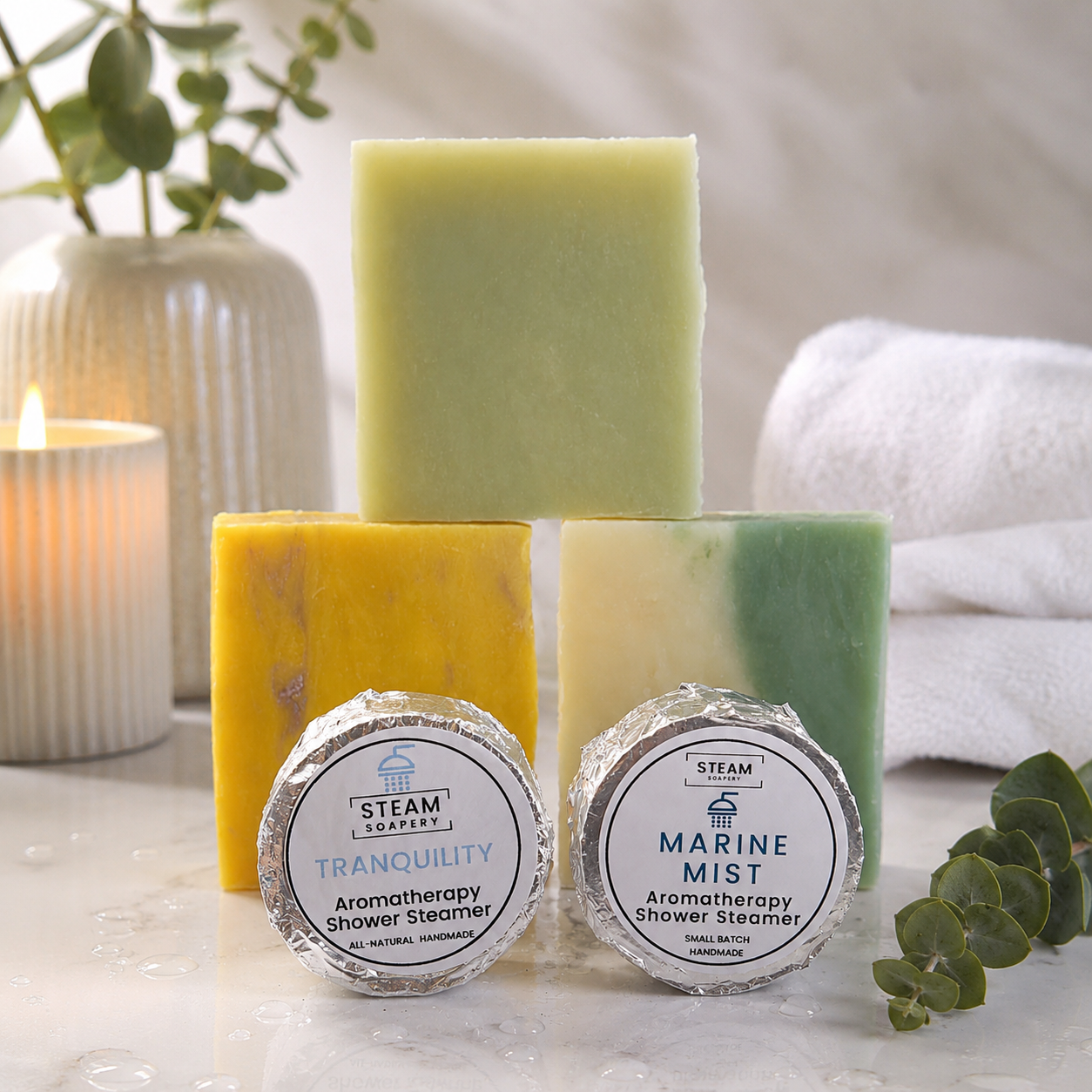 Three bars of soap with labels and two shower steamers on a marble surface with a candle and plant in the background.